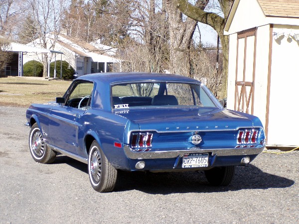 Blue 1967 Ford Mustang coupe parked on gravel, rear view. Houses and trees in background