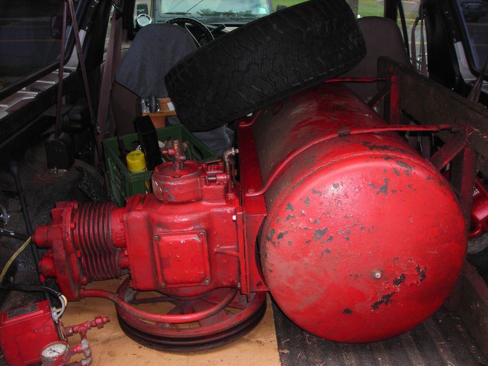 Air compressor and spare tire loaded in back of a vehicle. Some green crates and a yellow bottle are visible