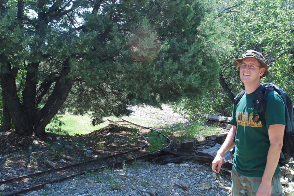 guy in green shirt and hat with backpack standing near some old train tracks and trees