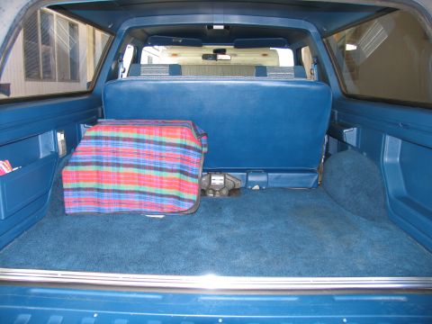 1984 Ford Bronco - The rear cargo area of a blue SUV features a plaid blanket covering an object and blue carpeting throughout.