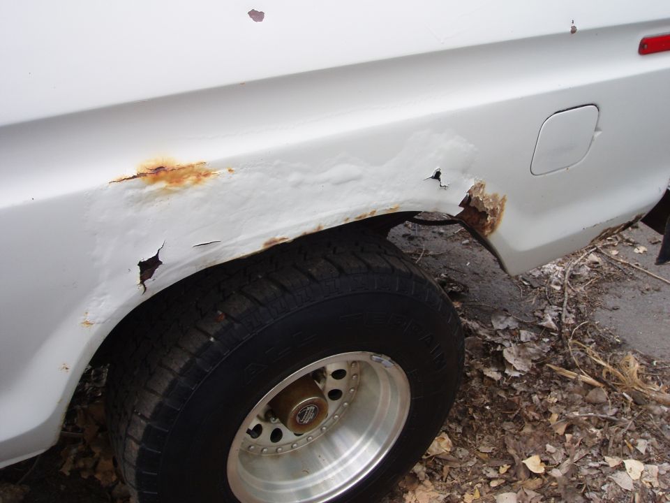 White vehicle's rear fender shows significant rust and holes. Tire and wheel are visible