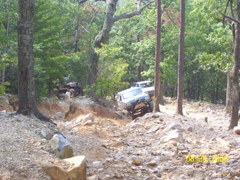 1978 Ford Bronco - Multiple off-road vehicles traverse a challenging rocky trail through a dense forest.