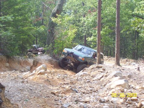 1978 Ford Bronco - Two off-road vehicles navigate a challenging, rocky trail through a wooded area.