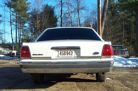 1991 Ford LTD Crown Victoria - Straight-on rear view of a white Ford Crown Victoria sedan on a dirt lot with snow patches and bare winter trees behind. The full-width chr…