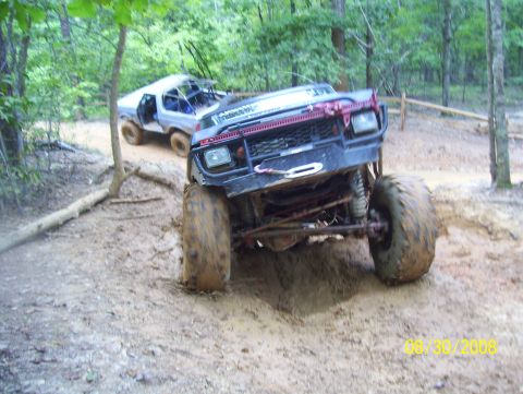 1978 Ford Bronco - Two off-road vehicles are stuck in the mud on a trail, surrounded by trees.