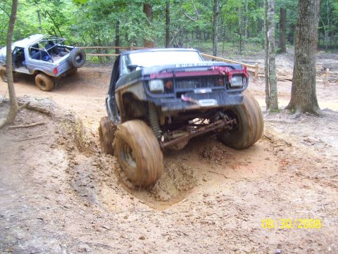 1978 Ford Bronco - Two modified off-road vehicles tackle a challenging muddy trail in a wooded area.