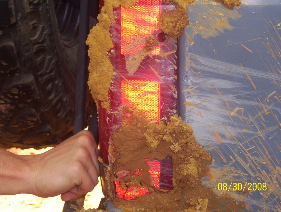 Mud covers a red taillight and part of a tire. A hand is visible near the taillight