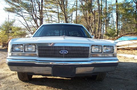 1991 Ford LTD Crown Victoria - Head-on front view of a white Ford Crown Victoria sedan parked on dirt in a wooded lot, showing the wide horizontal-bar grille with center…