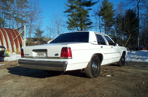 1991 Ford LTD Crown Victoria - Rear three-quarter view of a white Ford Crown Victoria four-door sedan parked on frozen dirt beside a rusted corrugated Quonset hut, with s…
