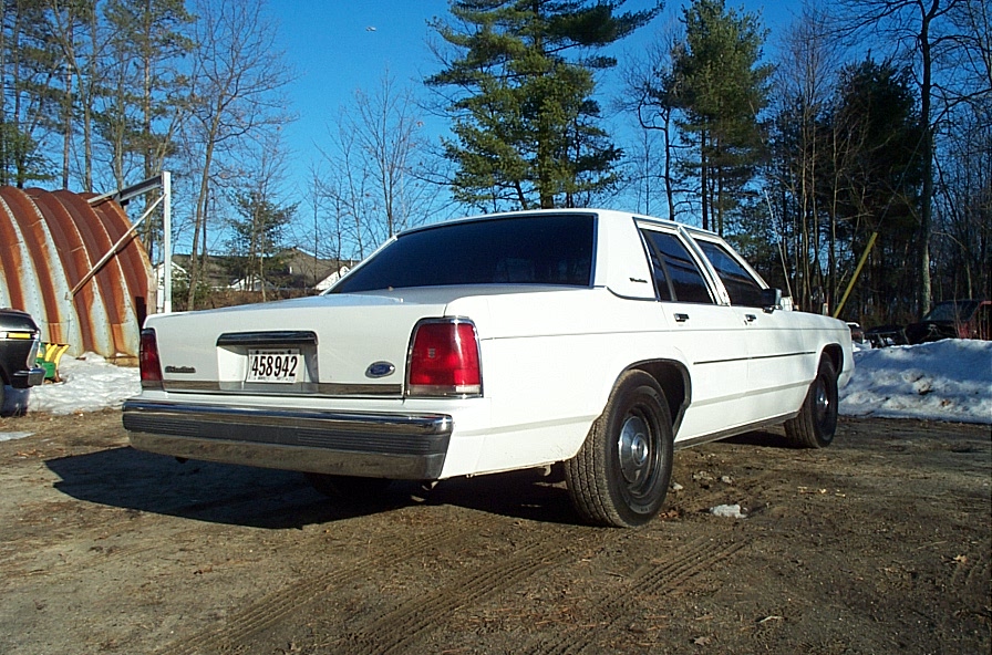 White Ford Crown Victoria sedan, rear 3/4 view on dirt lot with snow and Quonset hut