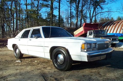 1991 Ford LTD Crown Victoria - Front three-quarter view of a white box-body Ford LTD Crown Victoria four-door sedan on a dirt lot, wearing black steel wheels with small c…