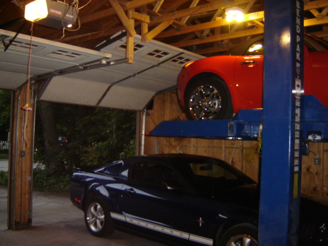 Dark blue Mustang on lift, red Mustang above it in garage