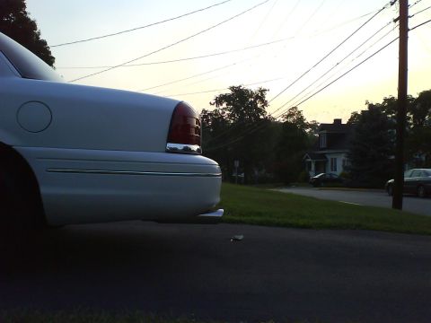 2003 Ford Crown Victoria - The rear of a white sedan is visible against a hazy sky, with a residential scene in the background.
