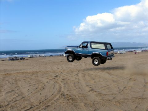 1984 Ford Bronco - A blue Ford Bronco is captured mid-air as it jumps over a sand dune at the beach.