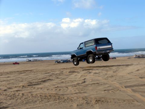 1984 Ford Bronco - A blue Ford Bronco is captured mid-air while jumping over sand dunes on a beach, with the ocean and other vehicles visible.