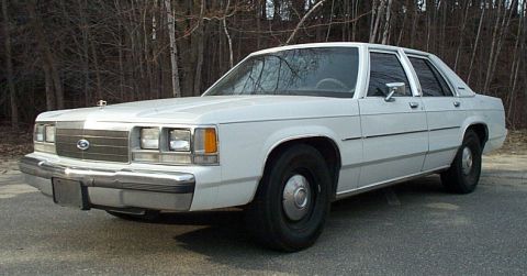 1991 Ford LTD Crown Victoria - White Ford Crown Victoria four-door sedan shot from the 3/4 front left on pavement in front of bare winter woods. It runs blackwall tires o…