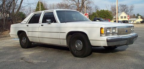 1991 Ford LTD Crown Victoria - White Ford Crown Victoria four-door sedan shot from the 3/4 front right in a parking lot, with a chrome pillar-mounted spotlight beside the…