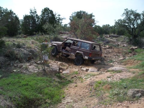 1996 Ford Bronco - A brown Jeep Cherokee XJ navigates a challenging rocky trail with its driver's door ajar.