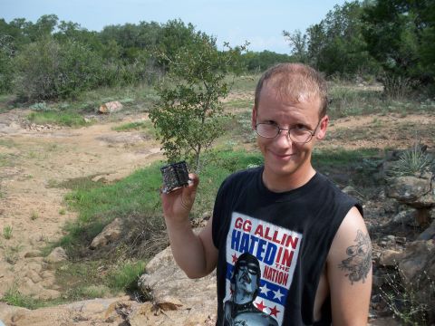 1996 Ford Bronco - A man wearing a GG Allin t-shirt holds a small, dark object while standing in a rocky, outdoor landscape.
