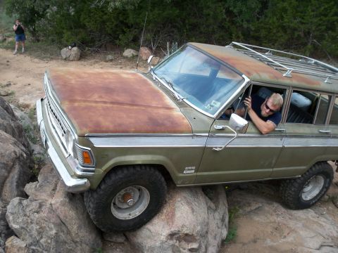1996 Ford Bronco - A man drives an olive green station wagon with a rusty hood over rocky terrain.