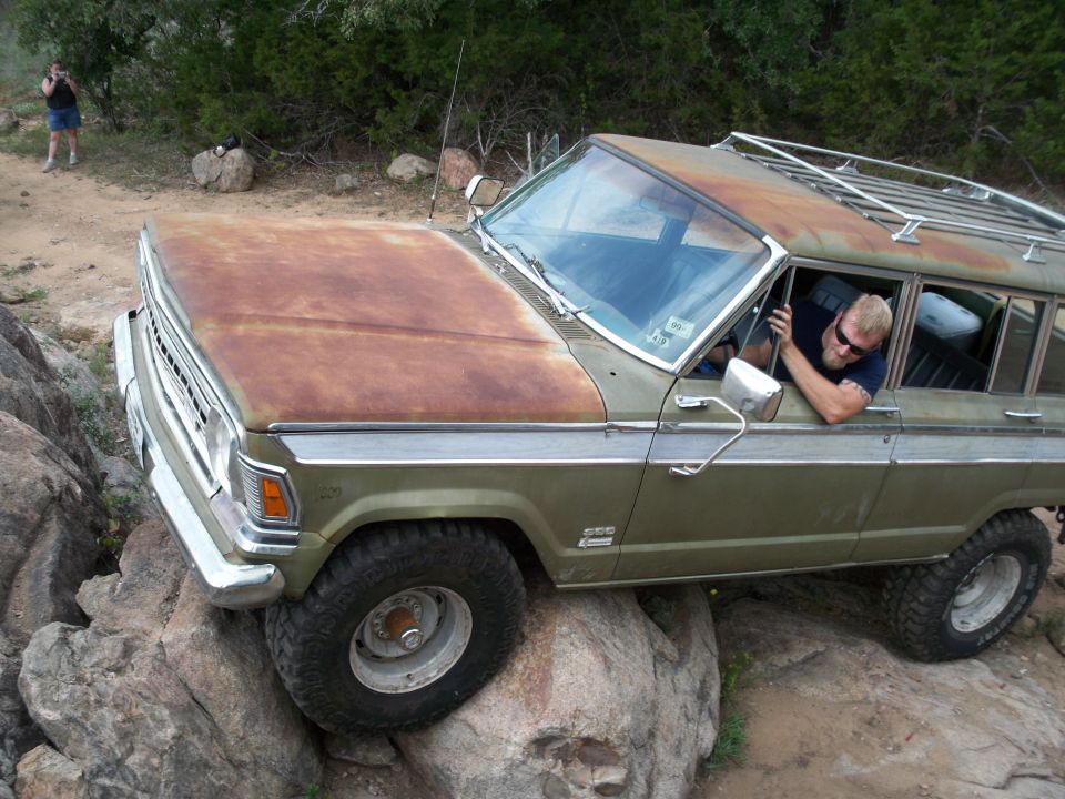 Olive green station wagon with rusty hood on rocks. Driver visible