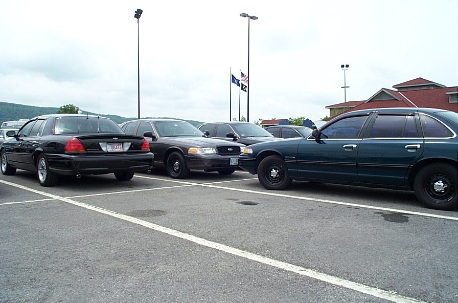 Three Ford Crown Victoria sedans parked in a lot. One is black, one is dark gray, and one is dark green