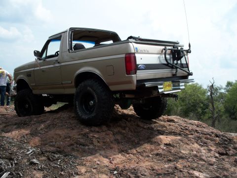 1996 Ford Bronco - A tan 1996 Ford Bronco is shown from the rear, equipped with off-road tires and a spare tire carrier, parked on rocky terrain.
