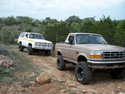 1996 Ford Bronco - Two lifted off-road vehicles, a Ford pickup and a Chevrolet Blazer, are parked on a dusty trail surrounded by trees.