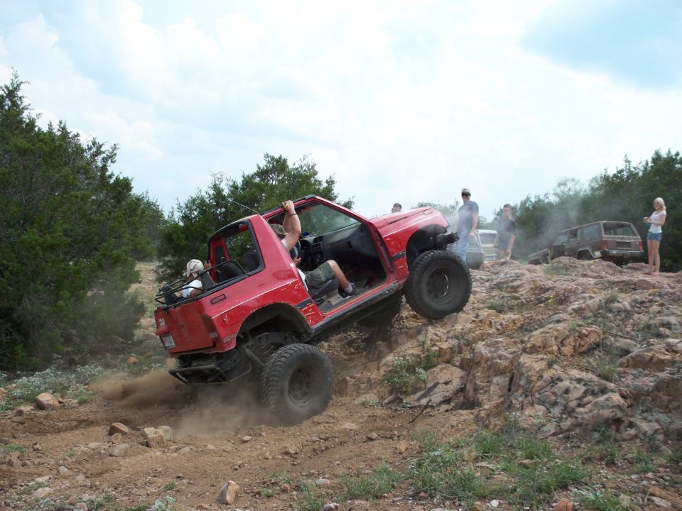 Red off-road vehicle with open doors climbing a rocky incline, kicking up dust. Other vehicles and people are visible in the background