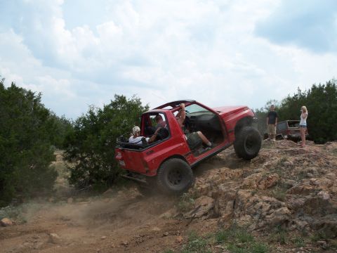 1996 Ford Bronco - A red off-road vehicle navigates a steep, rocky incline with passengers inside, while another vehicle and onlookers are visible in the back…