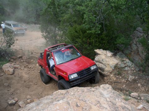 1996 Ford Bronco - Two SUVs navigate a rocky, off-road trail surrounded by trees and brush.