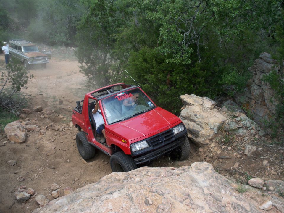 Red Suzuki Samurai off-roading on a dirt trail with rocks. Another older SUV is behind it