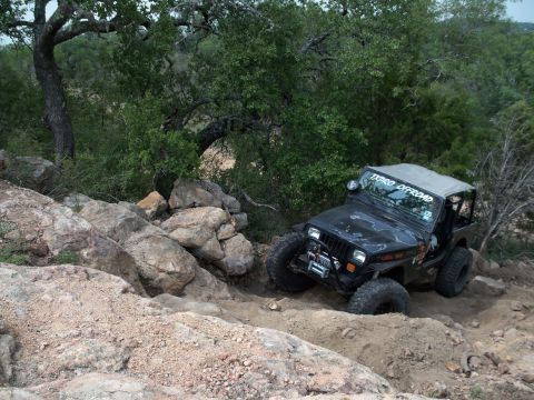 1996 Ford Bronco - A black Jeep Wrangler tackles a challenging off-road course with large rocks and dirt.