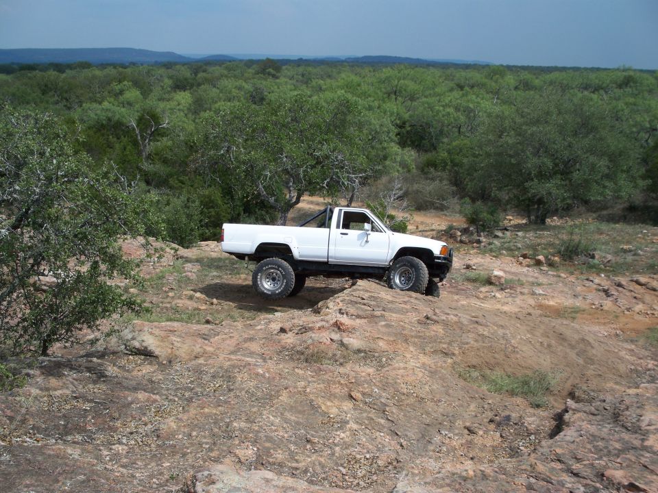 White pickup truck with off-road tires on a rocky incline, surrounded by trees