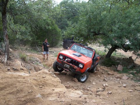 1996 Ford Bronco - An orange off-road vehicle is driving on a rocky, dirt trail surrounded by trees. Another vehicle is parked behind it.