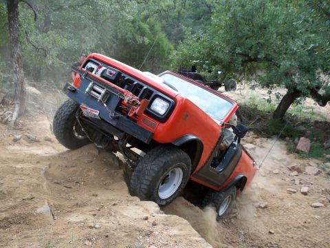 1996 Ford Bronco - An orange off-road vehicle navigates a challenging dirt trail, kicking up dust as it climbs.