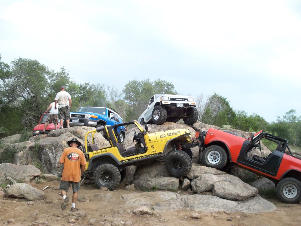 Several off-road vehicles, including a yellow Jeep, a red Jeep-like vehicle, a blue SUV, and a white pickup truck, are positioned on a rocky terrain