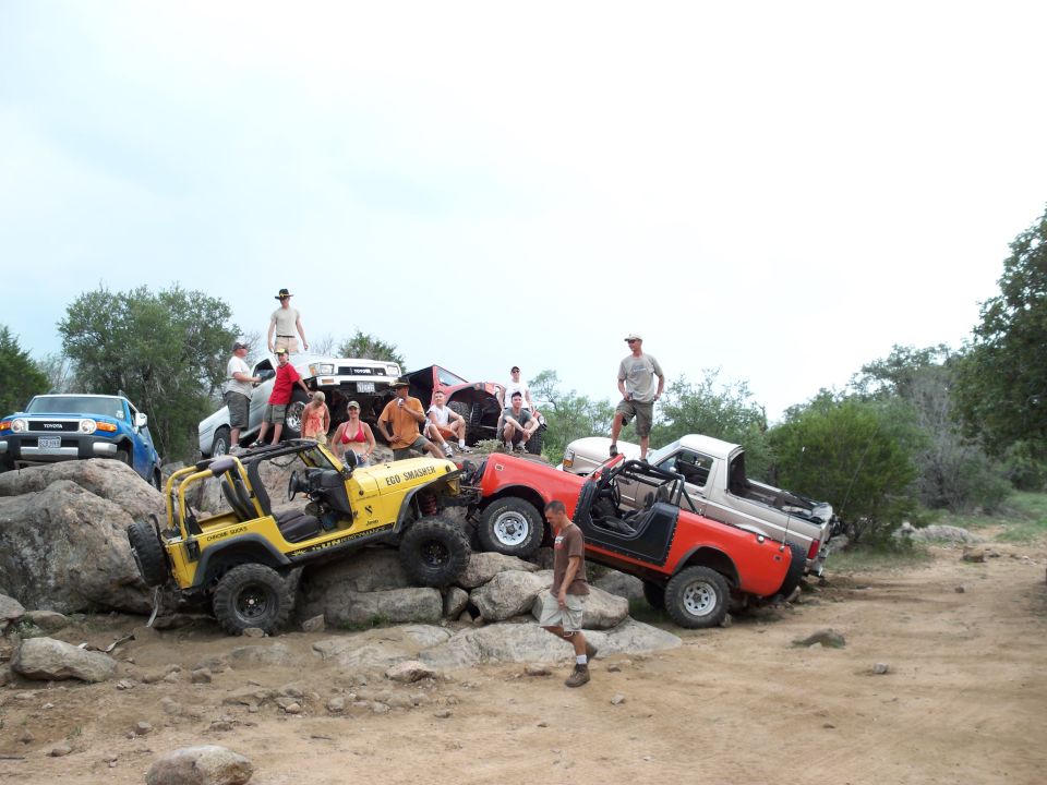 Several off-road vehicles, including a blue FJ Cruiser, yellow Jeep, red Jeep, and a pickup truck, are parked on rocks with people around them