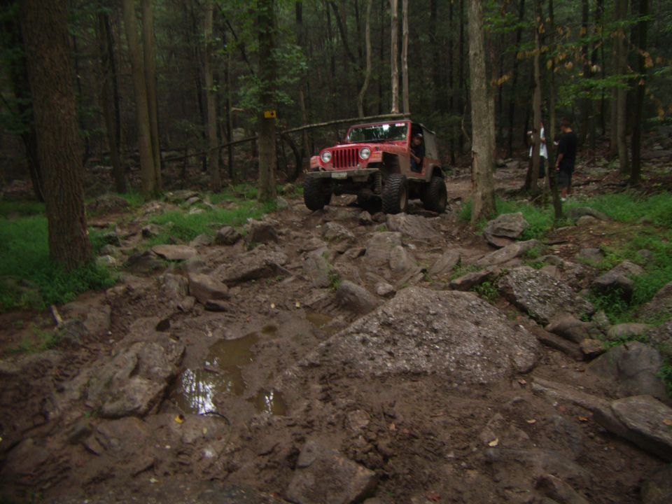 Red 2003 Jeep Wrangler on a muddy, rocky trail with trees and two people in the background