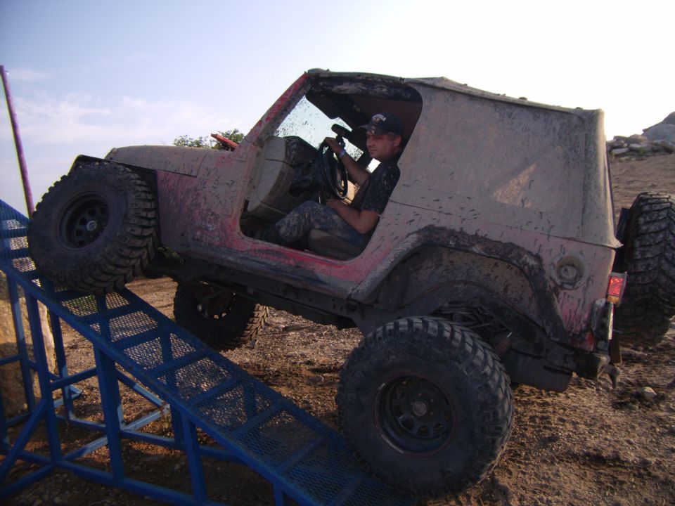 Mud-covered 2003 Jeep Wrangler driving up a blue metal ramp, driver visible inside