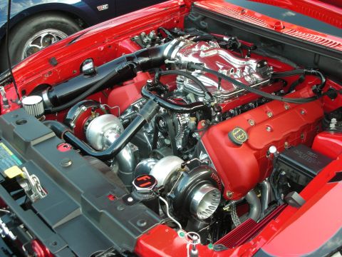 1998 Ford Crown Victoria - Close-up of a modified engine bay with twin turbochargers and chrome accents, partially obscuring another vehicle.
