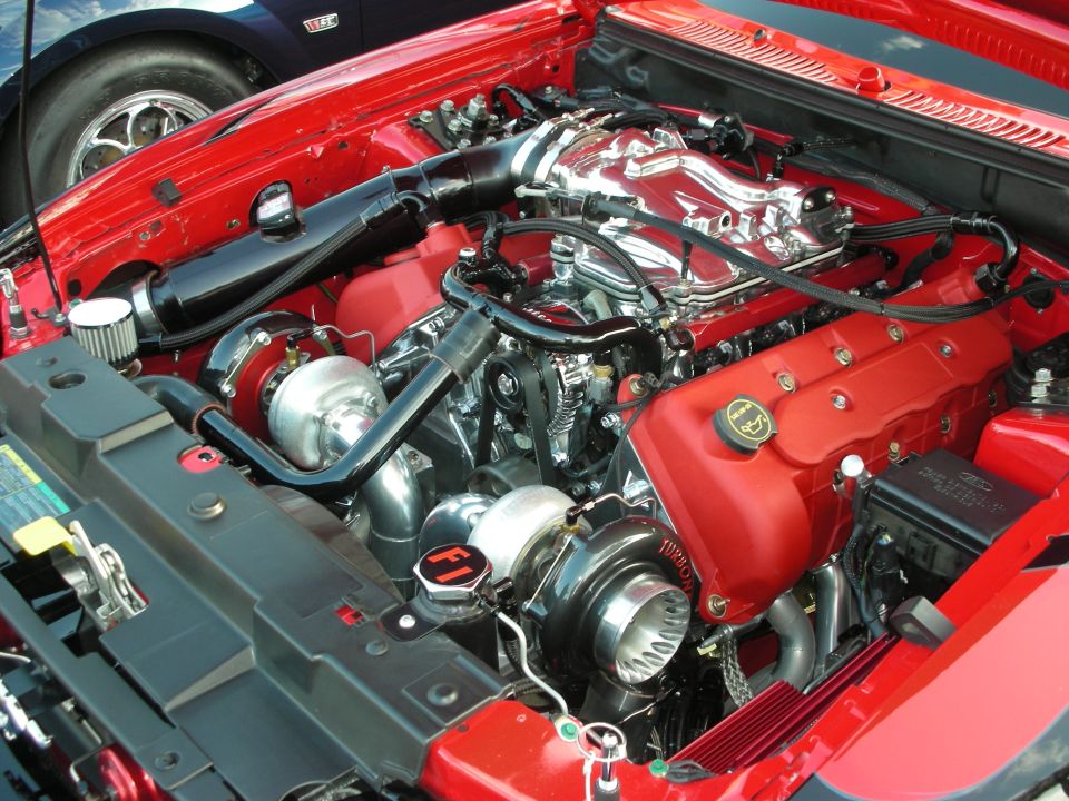 Red engine bay with twin turbochargers and chrome intake manifold. A dark blue car is partially visible in the background