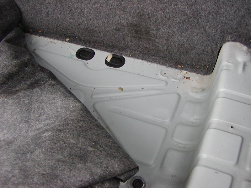 Car's rear passenger footwell area with carpet pulled back showing the metal floor pan and two rubber grommets