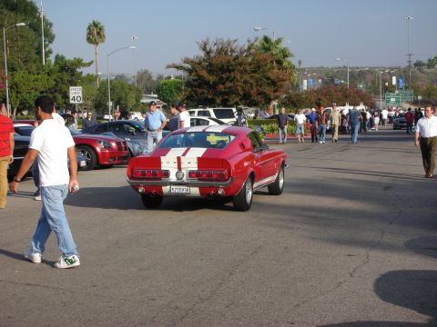 1998 Ford Crown Victoria - A red classic car with white racing stripes is parked at an outdoor gathering of vehicles and people.