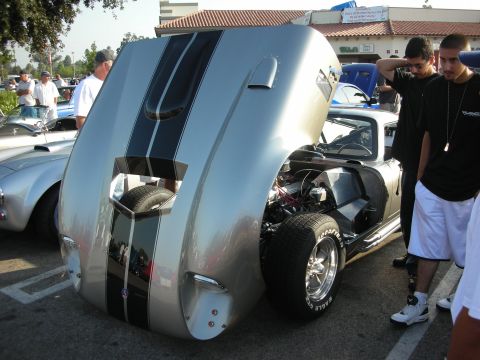 1998 Ford Crown Victoria - A silver sports car with dual black racing stripes has its hood open, displaying the engine and a spare tire. Another similar car is visibl…