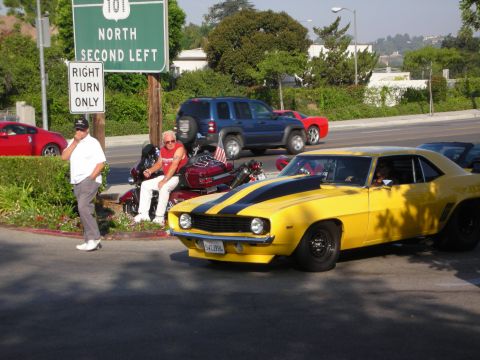 1998 Ford Crown Victoria - A yellow 1969 Chevrolet Camaro with black racing stripes is parked near a motorcycle and other vehicles.