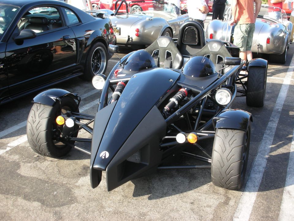Black Ariel Atom with two helmets in front of a black Mustang and a silver Cobra