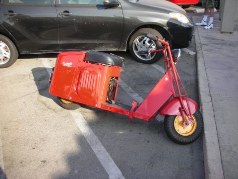 1998 Ford Crown Victoria - A red Cushman scooter is parked in a lot next to a dark-colored car.