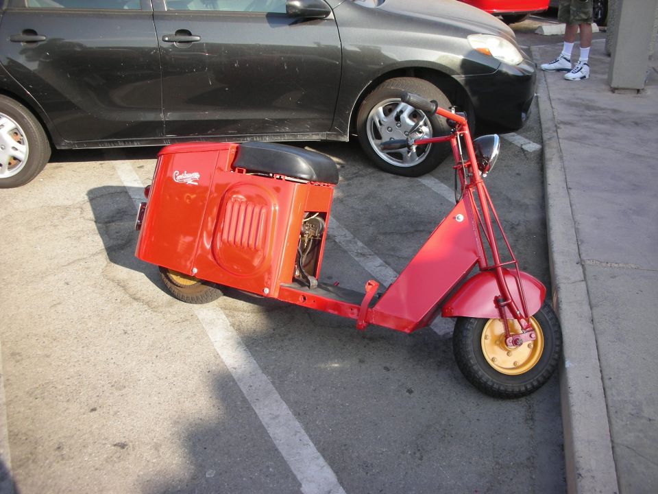 Red vintage scooter parked next to a black car. Text on the side reads 'Cushman'