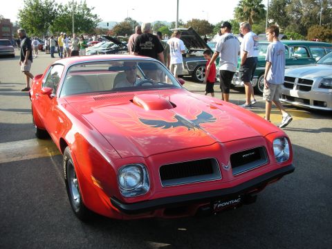 1998 Ford Crown Victoria - A red Pontiac Firebird with a distinctive hood scoop and flame decals is parked at an outdoor car meet.
